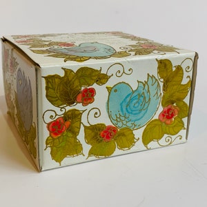 Puede incluir: Una caja blanca con un dise&ntilde;o floral azul y dorado. La caja presenta dos p&aacute;jaros azules con detalles dorados y flores rojas.