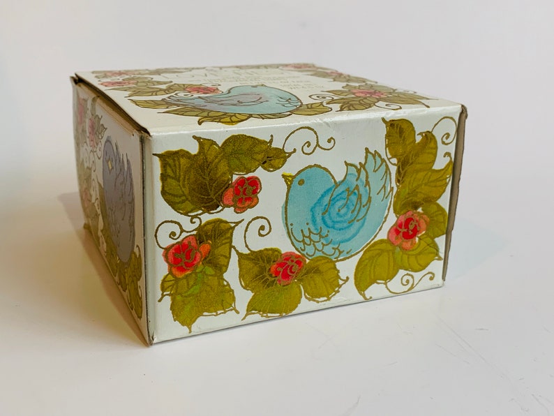 Puede incluir: Una caja blanca con un dise&ntilde;o floral azul y dorado. La caja presenta dos p&aacute;jaros azules con detalles dorados y flores rojas.