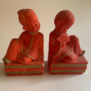May include: A pair of red bookends featuring two figures in traditional Chinese clothing. The figures are sitting on stacks of books, and one is holding a fan.