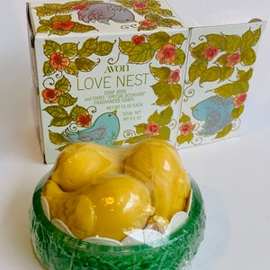 Puede incluir: Una caja verde y blanca con un dise&ntilde;o de p&aacute;jaro y el texto "AVON LOVE NEST SOAP DISH and THREE "SPECIAL OCCASION" FRAGRANCED SOAPS NET WT 1.5 OZ EACH TOTAL NET WT 4.5 OZ". La caja contiene tres jabones amarillos con forma de p&aacute;jaro en un nido verde.
