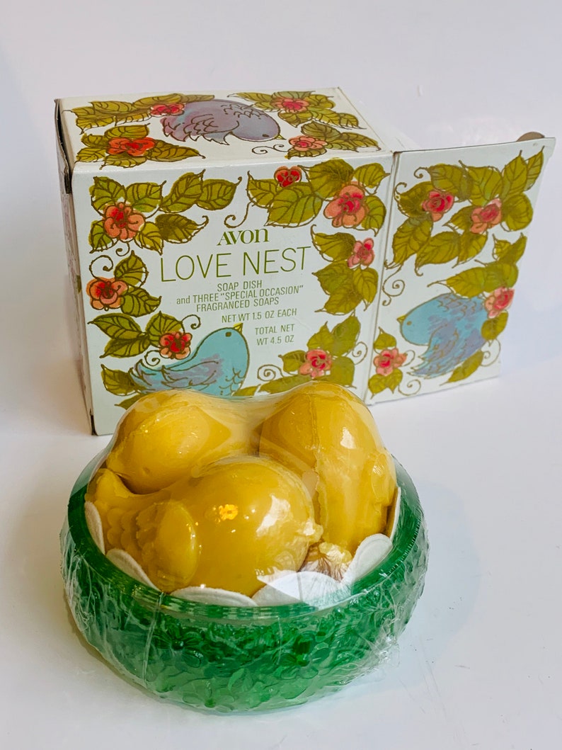 Puede incluir: Una caja verde y blanca con un dise&ntilde;o de p&aacute;jaro y el texto "AVON LOVE NEST SOAP DISH and THREE "SPECIAL OCCASION" FRAGRANCED SOAPS NET WT 1.5 OZ EACH TOTAL NET WT 4.5 OZ". La caja contiene tres jabones amarillos con forma de p&aacute;jaro en un nido verde.
