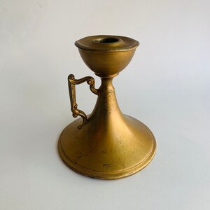 May include: A gold metal candle holder with a decorative handle. The holder has a round base and a flared top.
