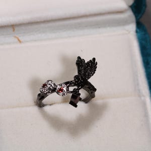 May include: A black ring with a bird design, set with red and clear gemstones. The ring is displayed in a white velvet jewelry box with a teal border. The bird has detailed wing features.