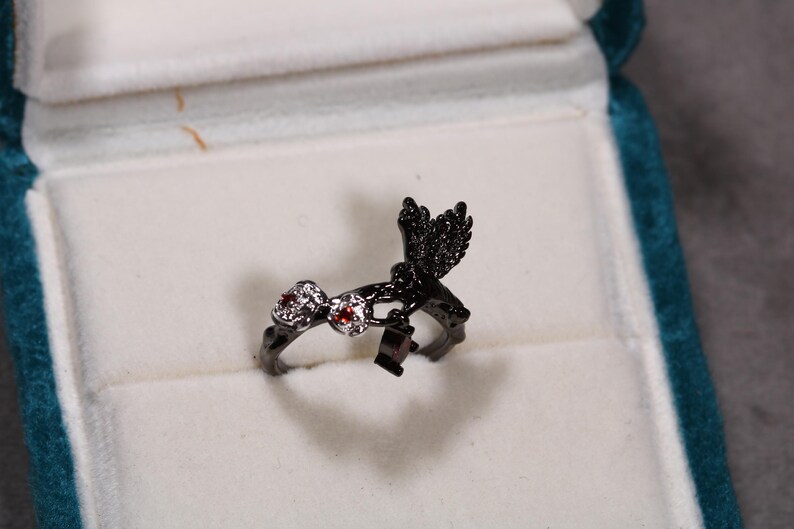 May include: A black ring with a bird design, set with red and clear gemstones. The ring is displayed in a white velvet jewelry box with a teal border. The bird has detailed wing features.