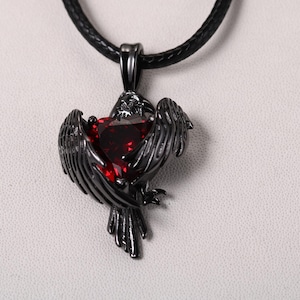 Puede incluir: Un collar con un colgante de metal negro y una piedra preciosa roja en el centro. El colgante tiene forma de cuervo con las alas extendidas.