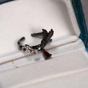May include: A black metal ring with a floral and bird design, set with small red gemstones. The ring is displayed in a teal velvet jewelry box, showcasing its intricate details and craftsmanship.