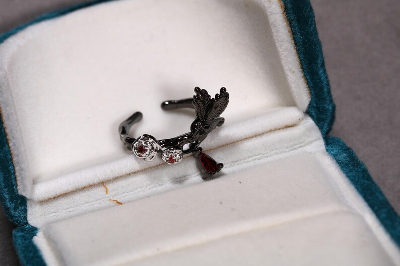 May include: A black metal ring with a floral and bird design, set with small red gemstones. The ring is displayed in a teal velvet jewelry box, showcasing its intricate details and craftsmanship.