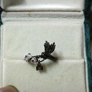 May include: A black ring featuring a bird perched on a branch with small white and red gemstones. The ring is set in a green velvet jewelry box with a white interior. The bird has detailed wing and tail feathers.