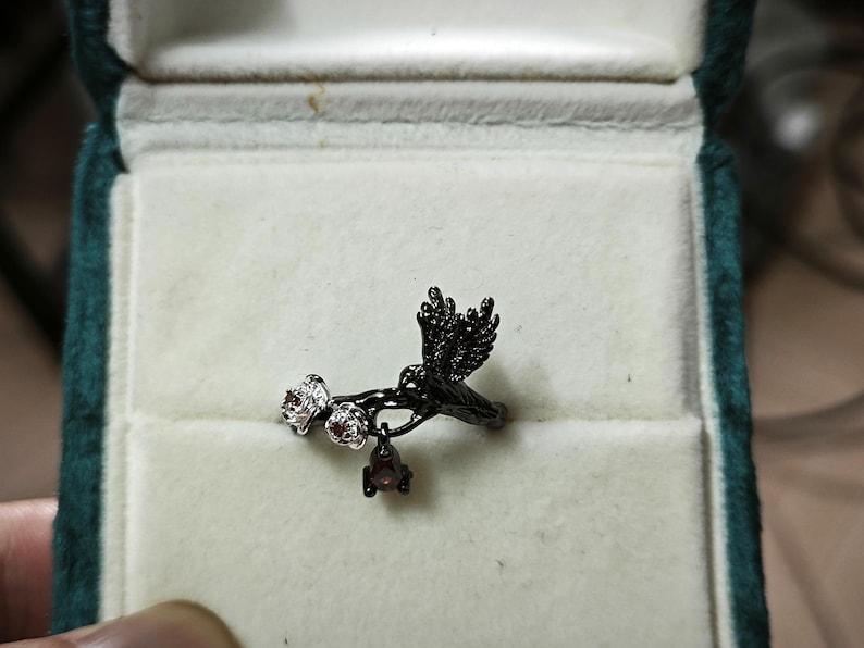 May include: A black ring featuring a bird perched on a branch with small white and red gemstones. The ring is set in a green velvet jewelry box with a white interior. The bird has detailed wing and tail feathers.