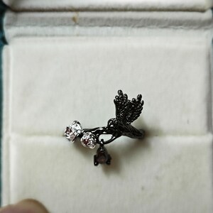 May include: Black metal ring featuring a bird in flight design with two small flower accents and a dangling gem. The ring is set against a white background inside a teal jewelry box.