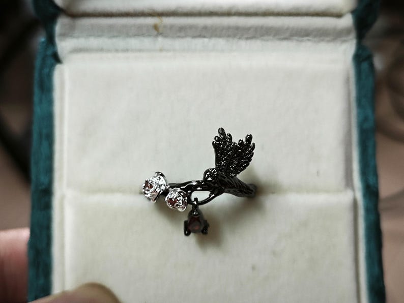 May include: Black metal ring featuring a bird in flight design with two small flower accents and a dangling gem. The ring is set against a white background inside a teal jewelry box.