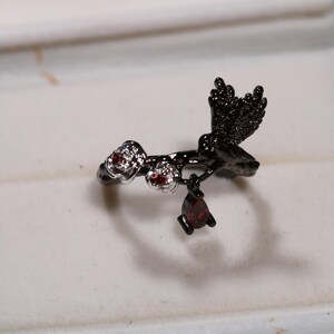 May include: A black metal ring featuring a hummingbird design with red gemstone accents. The ring has a branch-like band with a hummingbird perched on it, and a dangling teardrop-shaped gemstone.