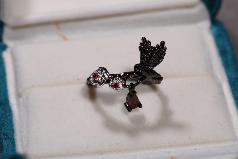 May include: A black metal ring featuring a hummingbird design with red gemstone accents. The ring has a branch-like band with a hummingbird perched on it, and a dangling teardrop-shaped gemstone.