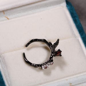 May include: Black ring with a branch-like design, featuring a red gemstone and two smaller clear gemstones. The ring is displayed in a teal jewelry box with a white interior.
