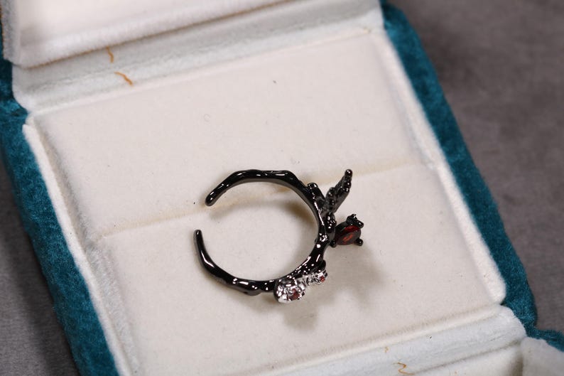 May include: Black ring with a branch-like design, featuring a red gemstone and two smaller clear gemstones. The ring is displayed in a teal jewelry box with a white interior.