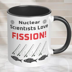 May include: A white ceramic mug with a black interior and handle. The front features the text "Nuclear Scientists Love FISSION!" in black and red, flanked by atom symbols. Below the text are five black line-art fishing rods and three fish.