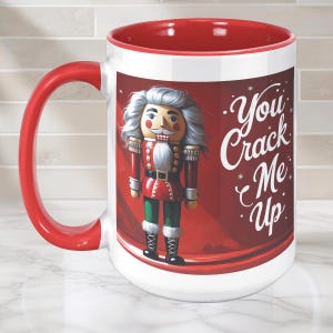 May include: A white ceramic mug with a red interior and handle, featuring a graphic of a nutcracker in a red and green uniform. The text "You Crack Me Up" is printed in white script on the right side of the mug against a dark red background.