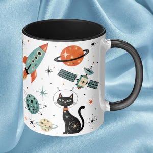 May include: White ceramic mug with a black interior and handle, featuring a retro space-themed design. The design includes a rocket, planets, a satellite, and a black cat in a space helmet. The mug is decorated with starbursts.