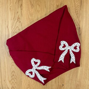 May include: A deep red hooded sweatshirt with two white and red polka-dot bow appliques. The bows have a heart-shaped center and green accents. The sweatshirt is folded on a wooden surface.