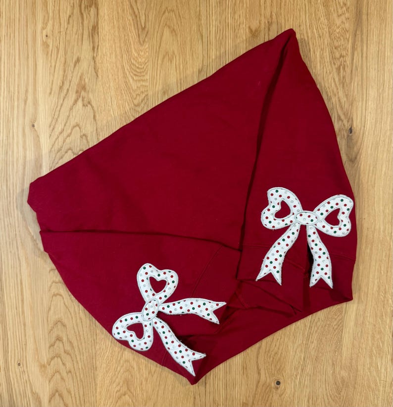May include: A deep red hooded sweatshirt with two white and red polka-dot bow appliques. The bows have a heart-shaped center and green accents. The sweatshirt is folded on a wooden surface.