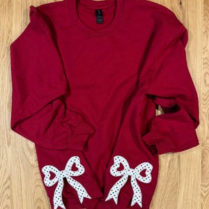 May include: A maroon sweatshirt with two white and polka-dotted bow appliques. The bows are positioned near the bottom hem of the long-sleeved top. The sweatshirt is made of a soft material and is suitable for casual wear.