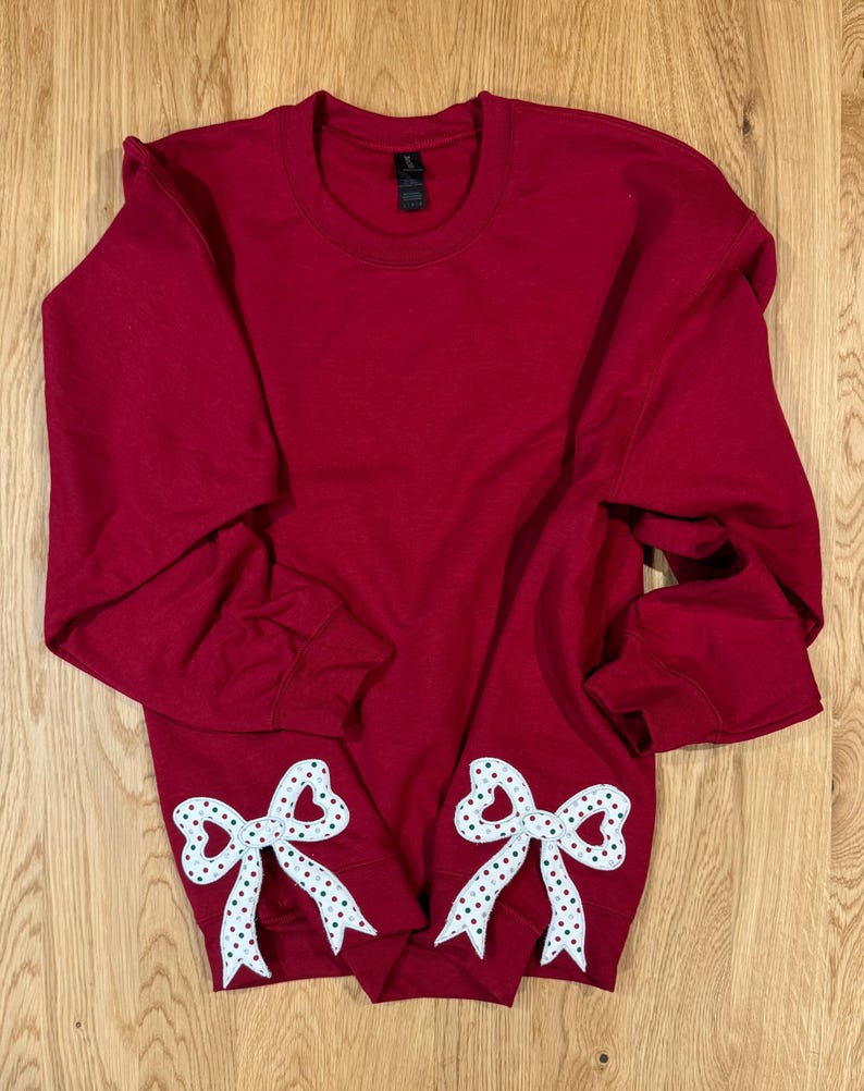 May include: A maroon sweatshirt with two white and polka-dotted bow appliques. The bows are positioned near the bottom hem of the long-sleeved top. The sweatshirt is made of a soft material and is suitable for casual wear.