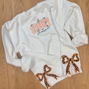 Puede incluir: Sudadera blanca con el texto "Spooky Season" en naranja y negro, con gráficos de fantasmas y murciélagos. Las mangas tienen lazos decorativos con un patrón de caramelos de Halloween. Una prenda festiva y acogedora.