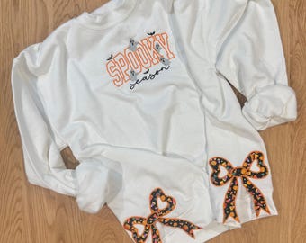 Puede incluir: Sudadera blanca con el texto "Spooky Season" en naranja y negro, con gráficos de fantasmas y murciélagos. Las mangas tienen lazos decorativos con un patrón de caramelos de Halloween. Una prenda festiva y acogedora.