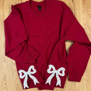 May include: A red sweatshirt with white bow appliques featuring red and green polka dots. The bows are placed on the lower front of the sweatshirt. The sweatshirt has a crew neck and long sleeves.