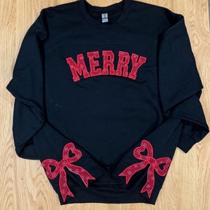 Pode incluir: Camisola de moletom preta com "MERRY" bordado em vermelho no peito e laços vermelhos com corações nas mangas.