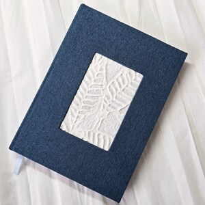 Peut inclure: Un journal en lin bleu foncé avec une fenêtre rectangulaire blanche présentant un motif de feuilles en relief.