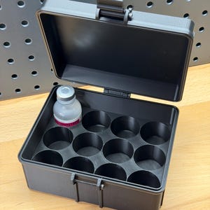May include: A black, rectangular storage case with a hinged lid, open to reveal a compartment with circular holders. A small bottle with a white cap and pink label is inside. The case is on a wooden surface.