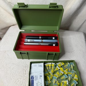 May include: An olive green plastic case with a red interior holds three insulin pens. A separate tray contains alcohol prep pads and yellow-capped needles. The case is open, revealing the contents.