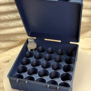 May include: A navy blue plastic storage case with a hinged lid and a grid of circular compartments. A small bottle sits in one of the compartments. The case is open, revealing the interior organization.