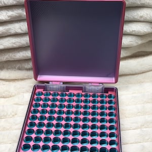 May include: A square storage case with a pink exterior and a gradient pink and teal interior. The case features a grid of circular compartments for small items, two hinges at the top, and two small silica gel packets placed inside the top row.
