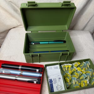 May include: An olive green plastic storage box with a hinged lid, open to reveal two pens. A red tray holds three pens, and a green tray contains alcohol prep pads and yellow-capped vials. The items are arranged on a white surface.