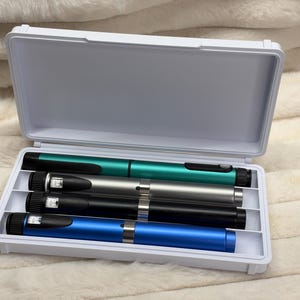 May include: A white plastic rectangular storage case containing four cylindrical injection pens. The pens are arranged horizontally in individual slots and feature metallic finishes in teal, silver, black, and blue, each with a black dial and window.