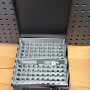 May include: Open black case with a gray interior, featuring multiple circular compartments. The case contains small items, including a vial and packets. The case is open, revealing the interior, and sits on a wooden surface.