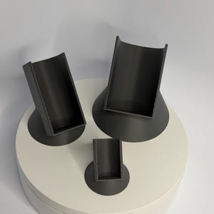 May include: Three dark gray, 3D-printed phone stands of varying sizes. Each stand has a curved, open-top design and sits on a circular base. The stands are arranged on a white, rotating platform.