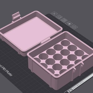 May include: A light pink, open rectangular container with a hinged lid. The interior of the container has multiple circular compartments. The lid is open, revealing the interior. The container is on a dark gray surface.
