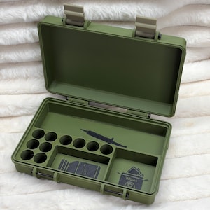 May include: An olive green rectangular storage case with a hinged lid. The interior features molded compartments, including circular slots, a long rectangular section with a syringe icon, and two square areas with icons for a bottle and wipes.