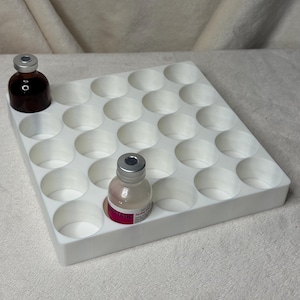 May include: A white square storage tray featuring twenty-five circular cutouts arranged in a five-by-five grid. Two small glass vials with metallic caps are placed in the tray, one containing a dark liquid and the other a light liquid.