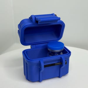 May include: A blue, open, small, rectangular container with a hinged lid. Inside the container is a small glass bottle with a blue cap. The container has a black latch and is sitting on a white surface.