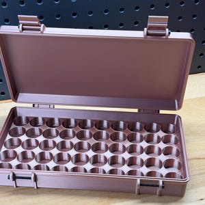 May include: Open, rose-colored case with a hinged lid and a tray filled with circular compartments. The case has a rectangular shape with a latch closure and is set on a wooden surface.