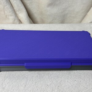 May include: A rectangular, closed storage box with a textured, royal blue lid and a gray base. The box has a handle on the front and is designed with reinforced corners. The box is sitting on a white surface.