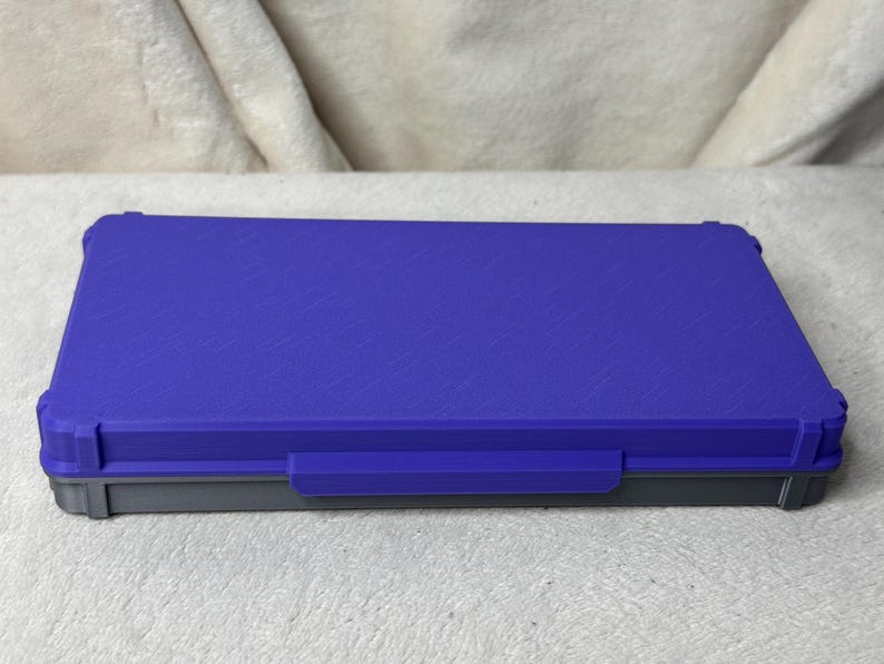 May include: A rectangular, closed storage box with a textured, royal blue lid and a gray base. The box has a handle on the front and is designed with reinforced corners. The box is sitting on a white surface.