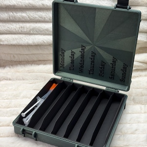 May include: An open, olive-green weekly pill organizer with black dividers and a hinged lid. The lid is labeled with the days of the week. Syringes are visible in one compartment. The case is designed to hold medications or medical supplies.