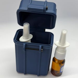 May include: A navy blue, rectangular case with a hinged lid, open to reveal a white nasal spray applicator. A second nasal spray bottle with a brown liquid sits beside the case. The case is designed to hold the nasal spray bottle.