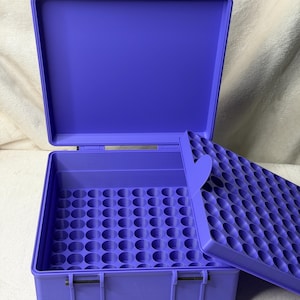 May include: A purple storage case with an open lid, revealing a grid of circular compartments. The case has a handle and latches for secure closure. The lid also has a grid of circular compartments. The case is designed for organizing small items.