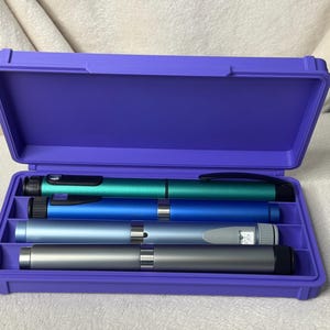 May include: A purple plastic case with an open lid, containing four insulin pens in shades of teal, blue, and silver. The pens are arranged neatly within the case, which is set against a neutral background.